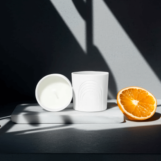 Composition sur marbre blanc : deux pots a bougie blancs (un debout motif arches, un couche cire blanche). A droite, une demi-orange juteuse. Ombres geometriques dramatiques traversent la scene, creant un contraste saisissant.