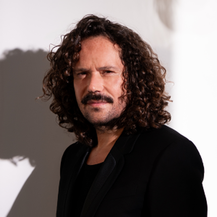 Portrait d'un homme au regard direct, avec des cheveux boucles fonces et une moustache. Il porte une veste noire, il a un style decontracte et elegant. L'eclairage met en valeur son visage sur un fond clair avec des jeux d'ombres subtils.