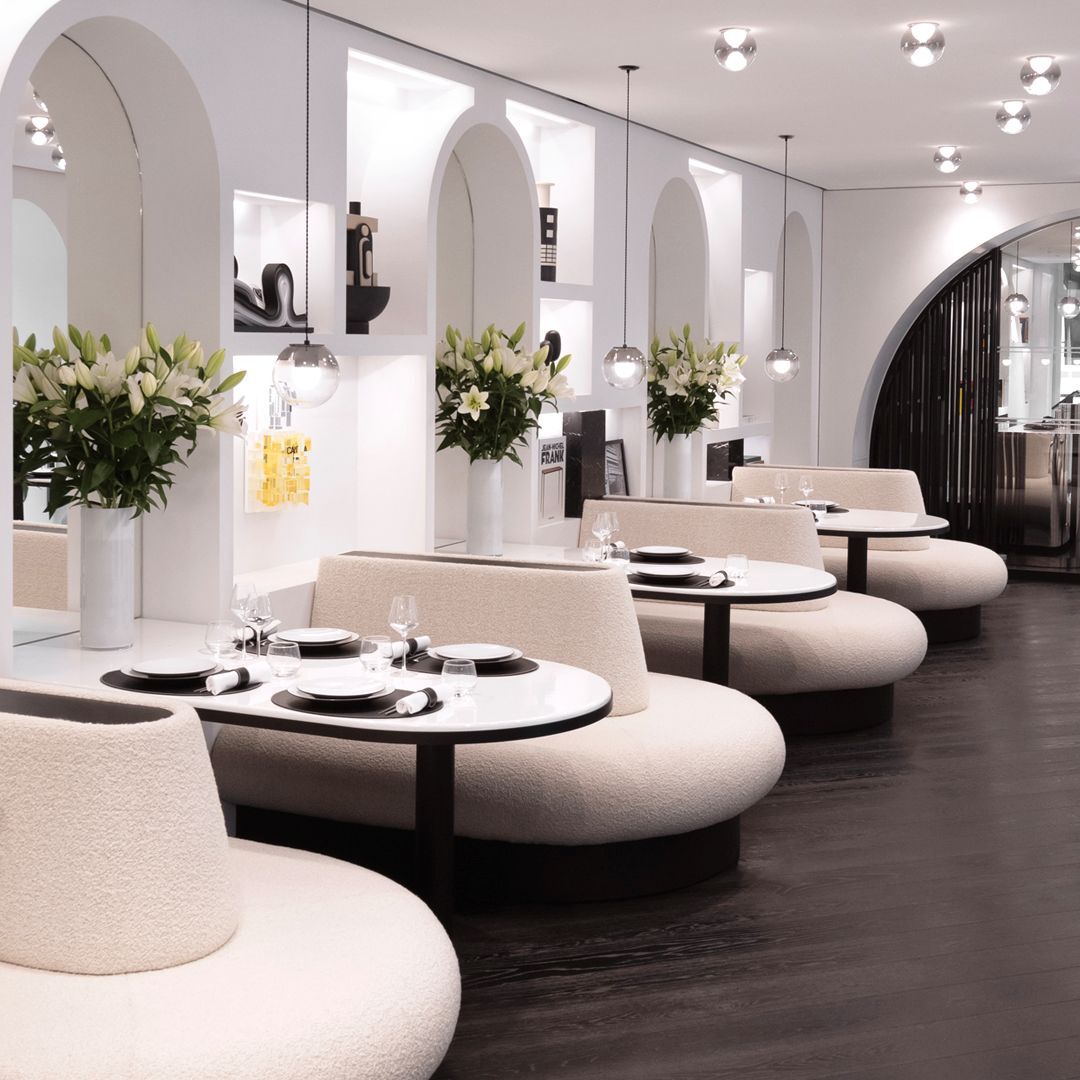 Espace repas elegant avec banquettes courbees, tables rondes dressees, murs blancs arques avec alcoves, et bouquets de lys blancs. Ambiance chic et moderne.