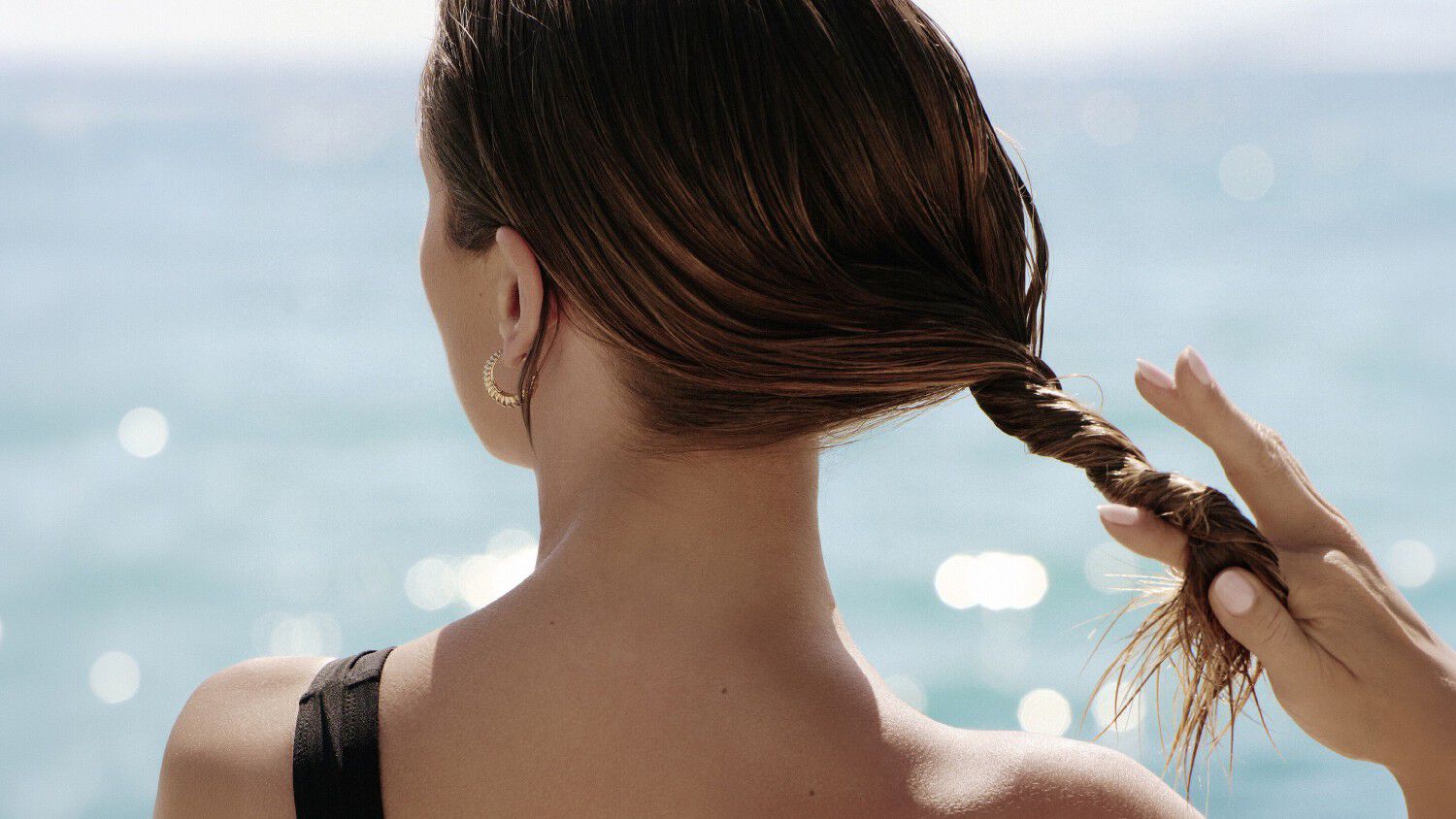 Femme de dos au bord de l'eau passant ses mains dans ses cheveux.