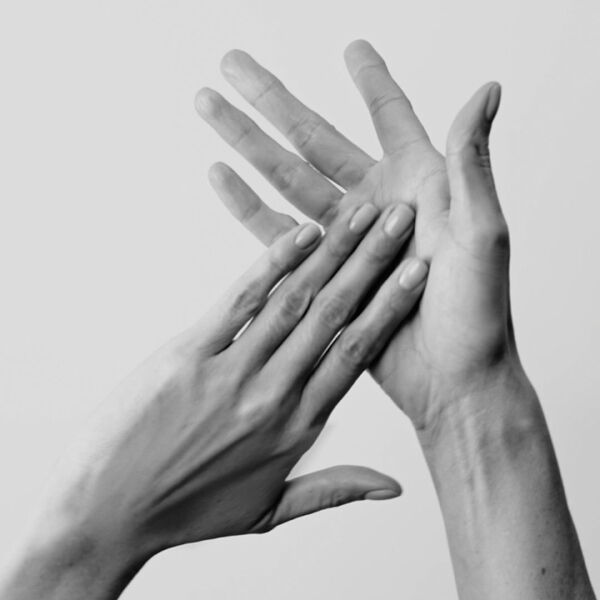 Gros plan monochrome sur des mains en mouvement, l'une caressant doucement l'autre, evoquant un soin ou une application de produit, avec une manucure soignee.