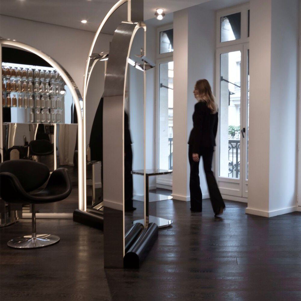 Un salon de coiffure moderne et elegant. De grandes structures arquees reflechissantes integrent l'eclairage. Un fauteuil noir design est a gauche. Des etageres murales affichent des flacons. Une femme s'eloigne vers des fenetres avec balcon. 