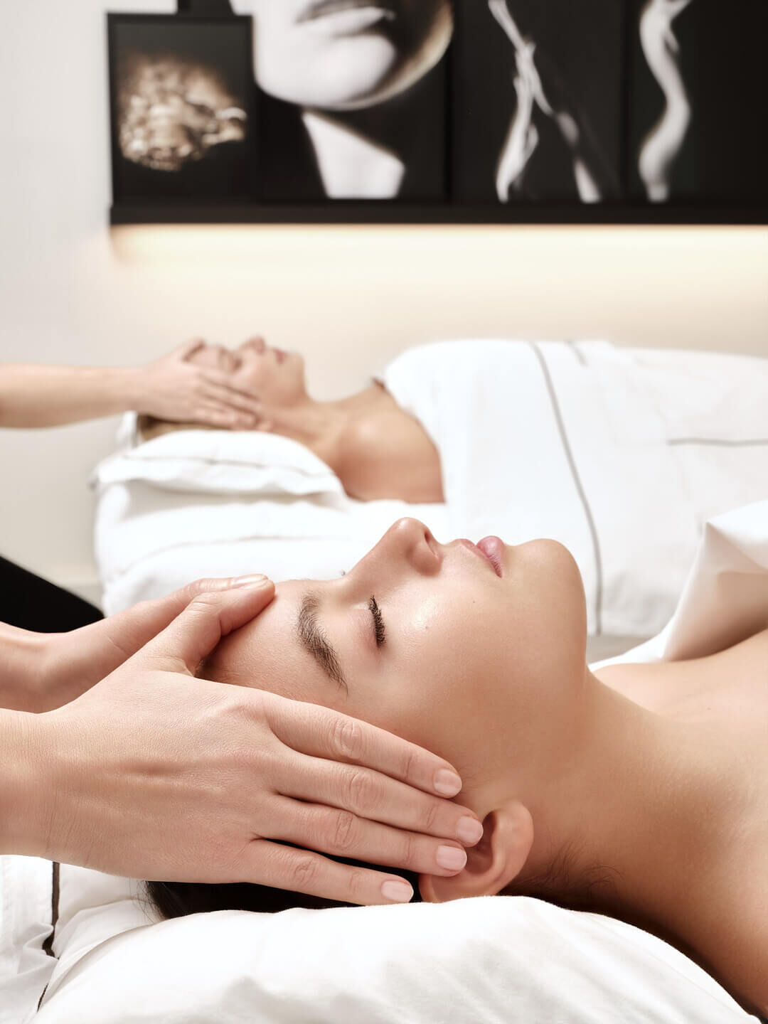 Deux femmes se detendent, yeux fermes, recevant de doux massages tete et visage en spa, evoquant la serenite et le bien-etre.