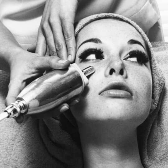 Une femme recevant un soin esthetique, avec un appareil sur son visage, tandis qu'une main l'examine.