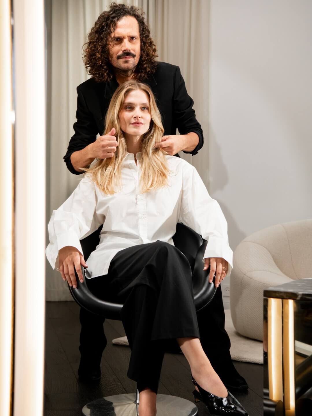 Un maitre styliste, d'un geste assure, parfait la chevelure blonde lumineuse d'une cliente, incarnant le luxe sur mesure et l'artisanat d'exception du salon.