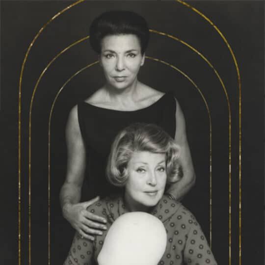 Deux femmes posent ensemble sur un fond noir, l'une debout derriere l'autre, avec une lumiere douce accentuant leurs visages.