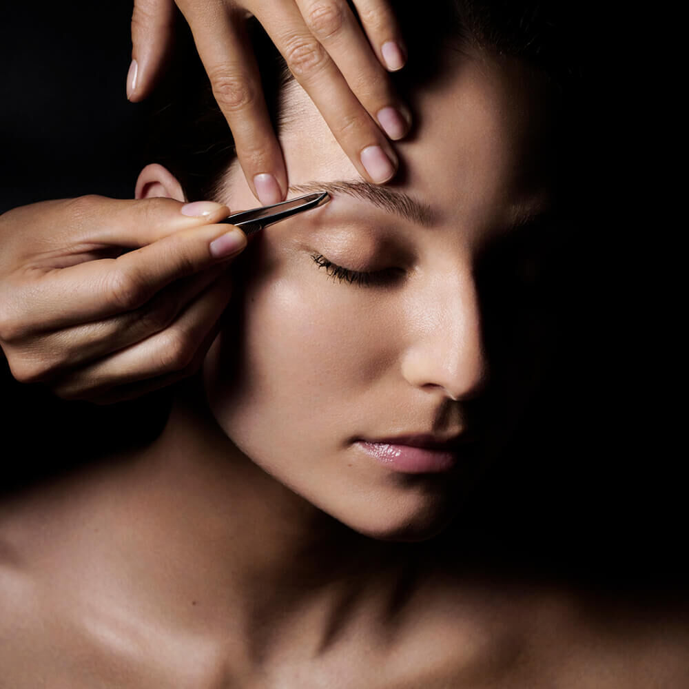 Gros plan sur le visage d'une personne qui a un oeil ferme, tandis qu'une main utilise une pince a epiler pour dessiner son sourcil sur un fond sombre.