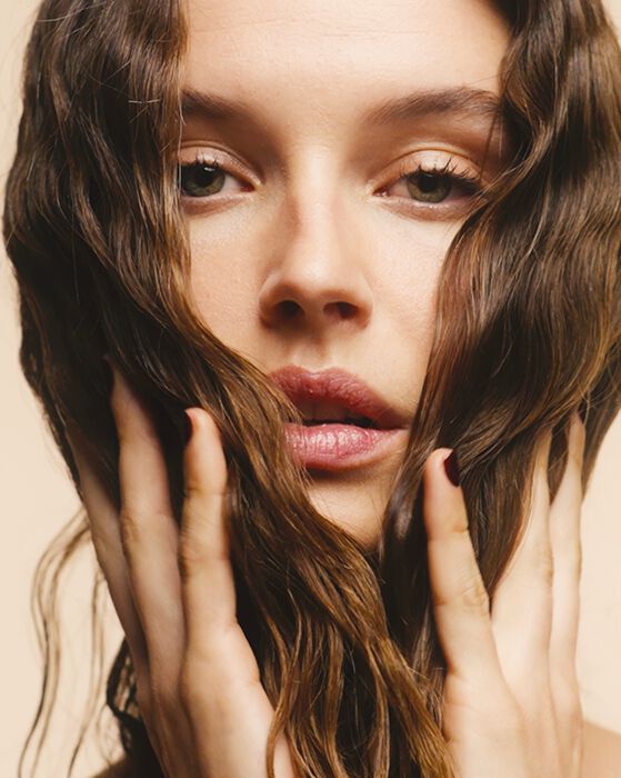 Gros plan d'une femme aux cheveux bruns ondules, encadrant son visage avec ses mains, sur un fond beige clair.