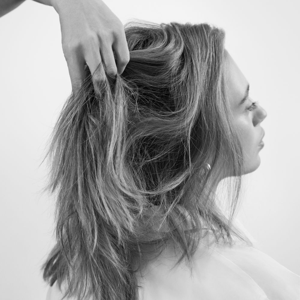 Une image en noir et blanc montre le profil de la tete d'une personne, tandis qu'une main souleve delicatement ses cheveux mi-longs, ondules et volumineux, mettant en valeur leur texture et leur mouvement.