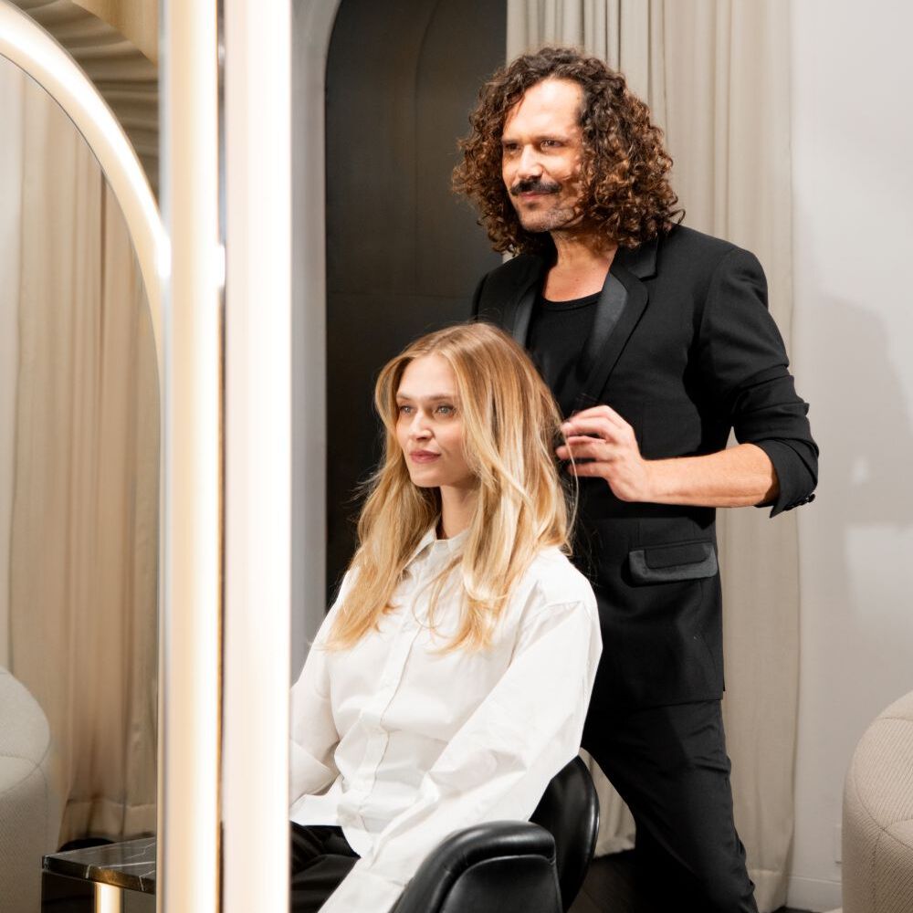 Dans un salon moderne, une femme blonde en chemise blanche est assise dans un fauteuil noir, regardant a gauche. Un homme en costume noir ajuste sa coiffure. La scene est encadree par des miroirs, refletant des rideaux beiges et une arche sombre.