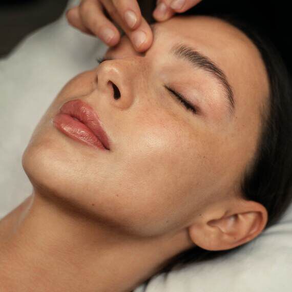 Gros plan du visage d'une personne aux yeux fermes recevant un doux massage facial sur le front, revelant une texture de peau naturelle et une expression paisible.