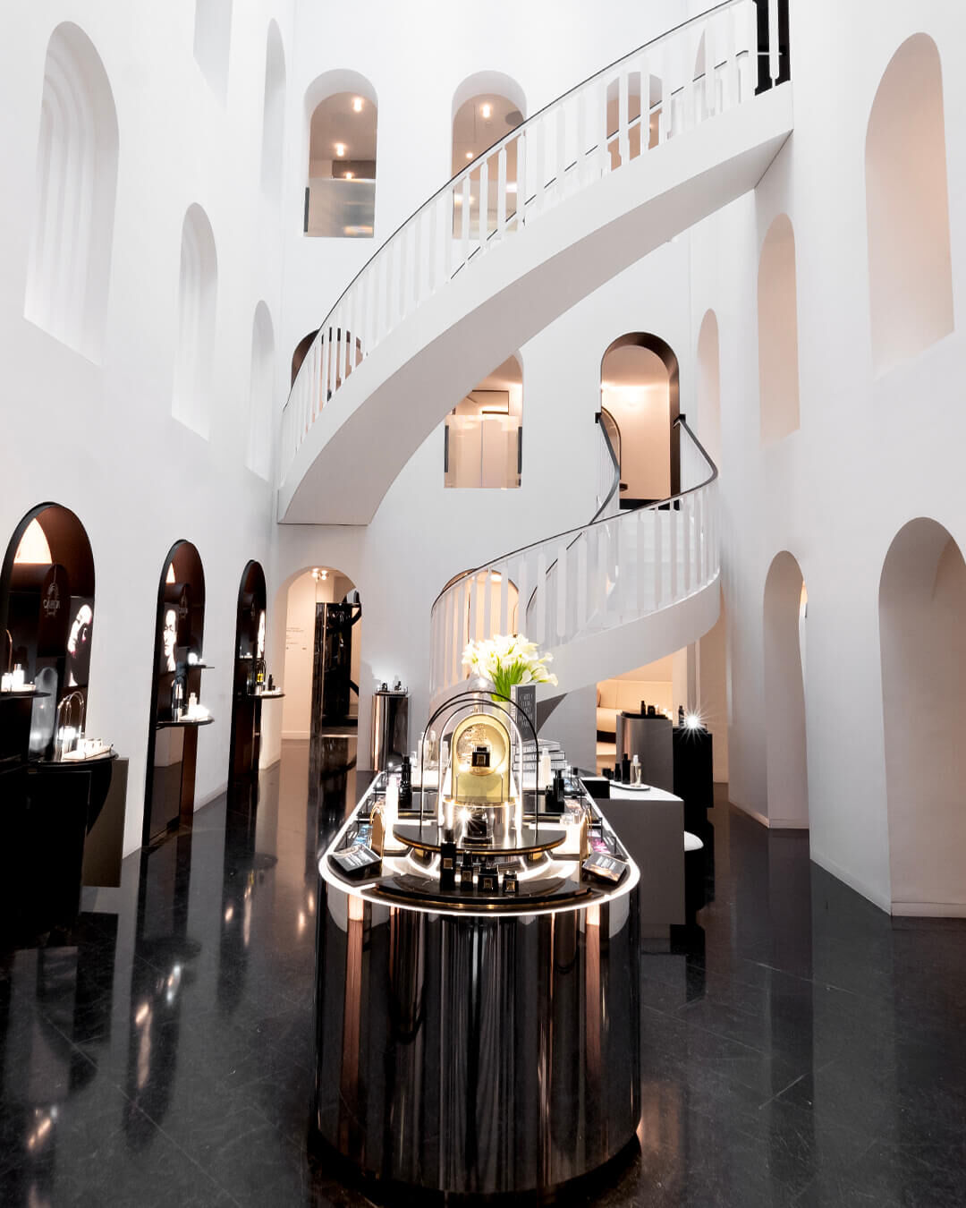 Interieur moderne et luxueux d'une boutique avec grand escalier blanc en colimacon, arches, et presentoirs de produits sur sol noir brillant.