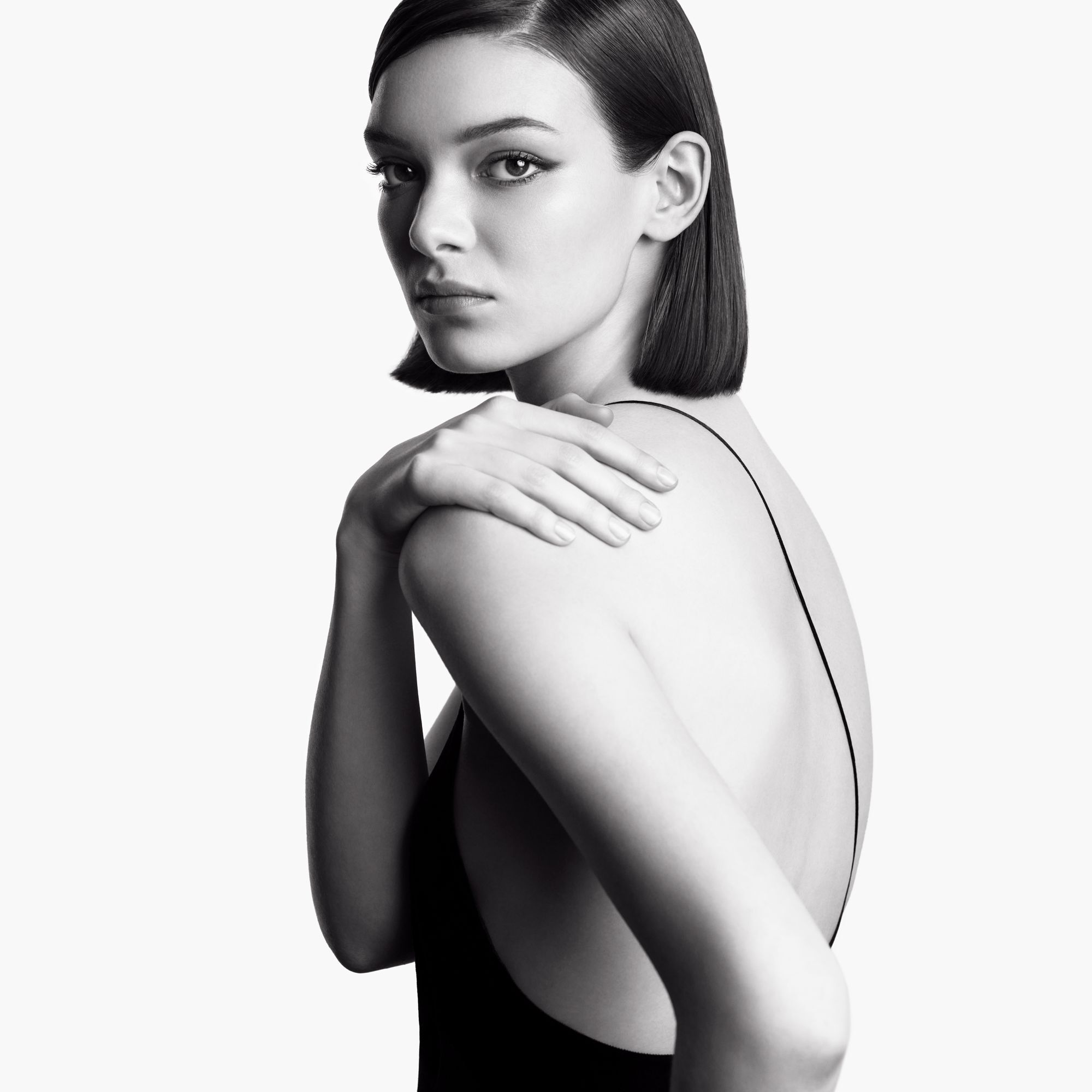 Portrait monochrome d'une femme aux cheveux courts, posant dos a la camera et regardant par-dessus son epaule, avec une expression intense et sophistiquee. 