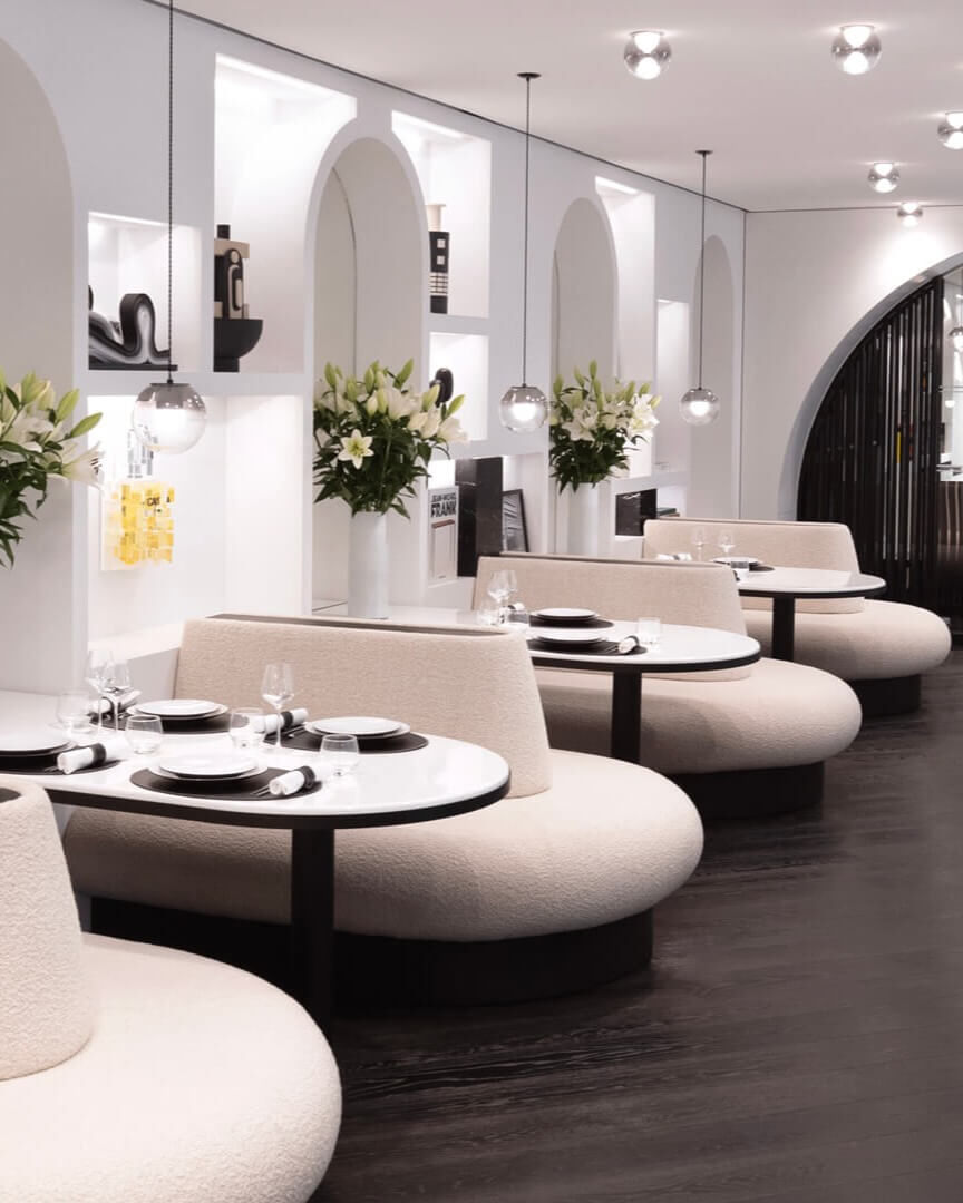 Espace repas elegant avec banquettes courbees, tables rondes dressees, murs blancs arques avec alcoves, et bouquets de lys blancs. Ambiance chic et moderne.