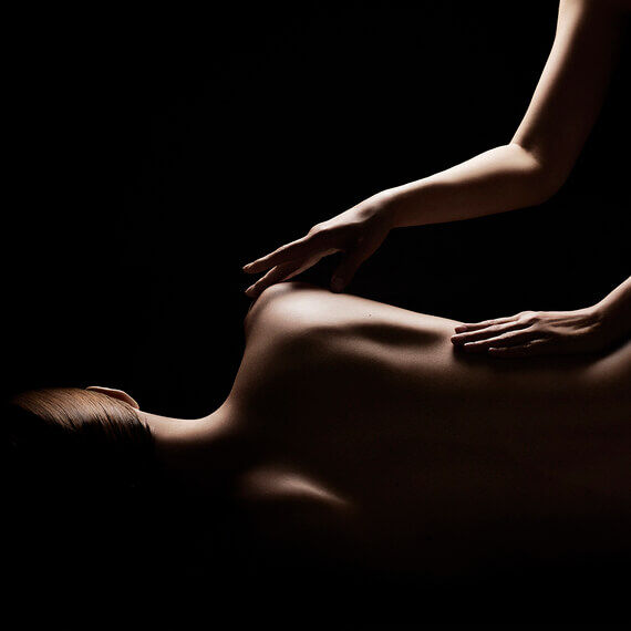 Deux mains effleurent delicatement le dos nu d'une personne allongee, mise en lumiere subtilement sur un fond sombre, suggerant detente et soin.