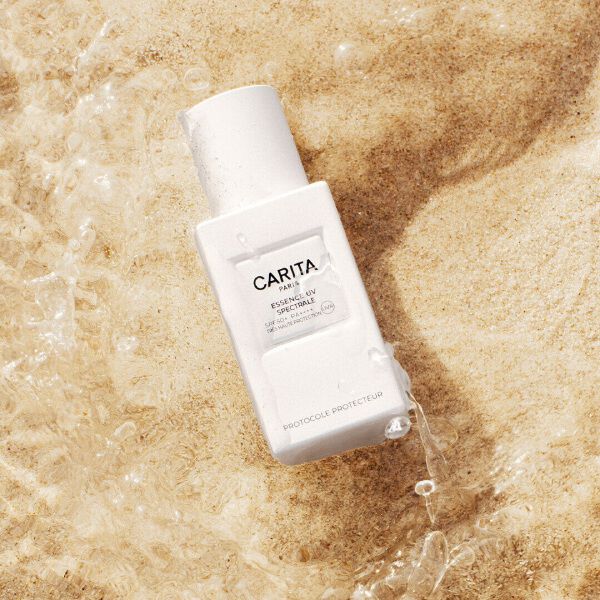 Flacon blanc de l'Essence UV de Carita pose dans le sable.