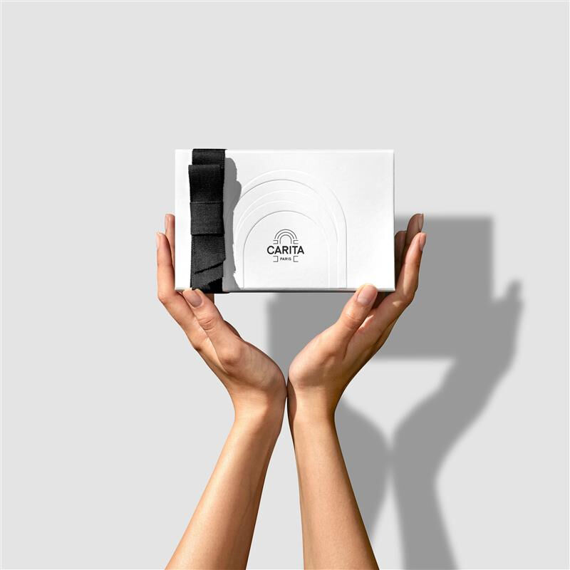 Les mains d'une femme tenant un coffret cadeau de luxe Carita Paris blanc, orne d'un elegant noeud en ruban noir.