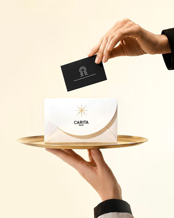 Des mains presentant une carte cadeau Carita Paris et une enveloppe blanche sur un plateau dore. La presentation luxueuse evoque un service haut de gamme et une experience beaute soigneusement selectionnee.