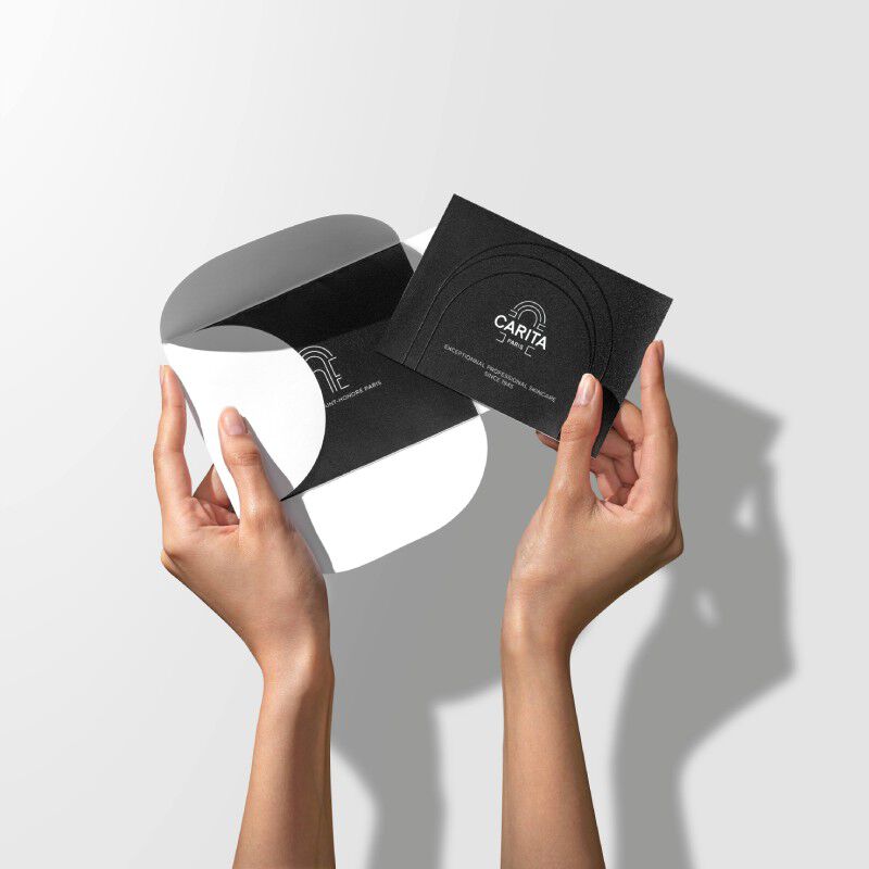 Des mains tenant une carte Carita Paris noire minimaliste et une enveloppe graphique noire et blanche sur un fond neutre.