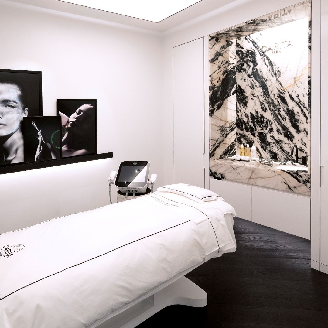 Salle de soin moderne et luxueuse avec un lit de traitement blanc, un appareil esthetique, et un decor mural en marbre noir et blanc, complete par des photos.