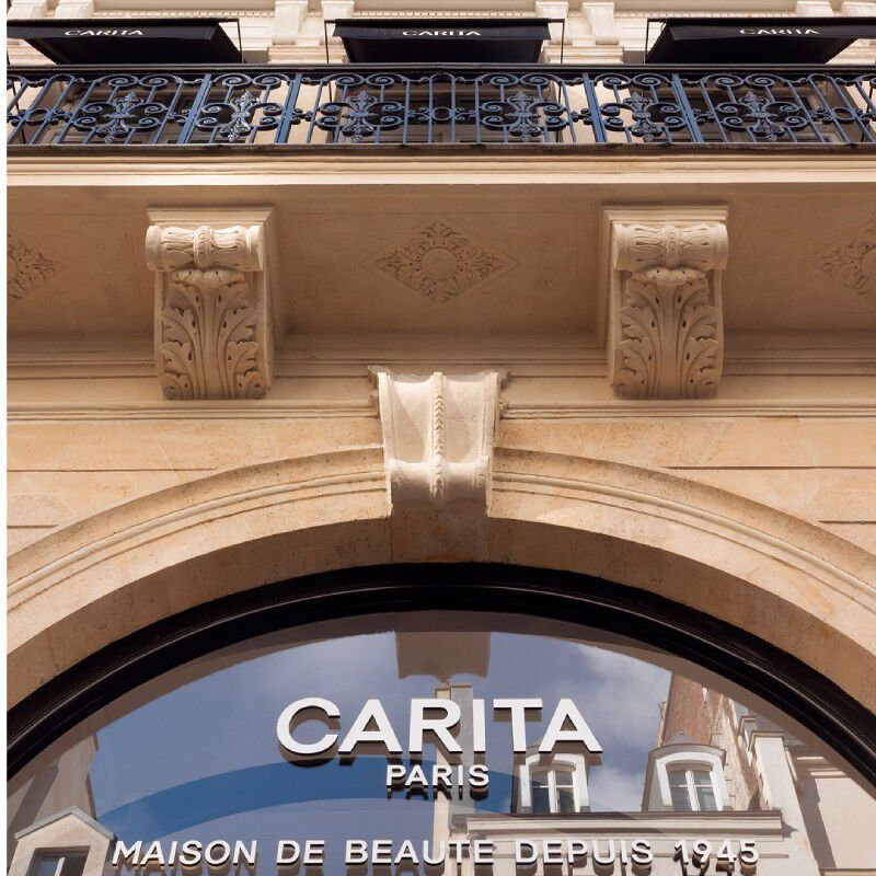 Facade elegante de la Maison Carita Paris. On voit un balcon en fer forge, des details architecturaux sculptes et le logo Carita Paris sur la vitrine incurvee. Marque "Maison de Beaute Depuis 1945".